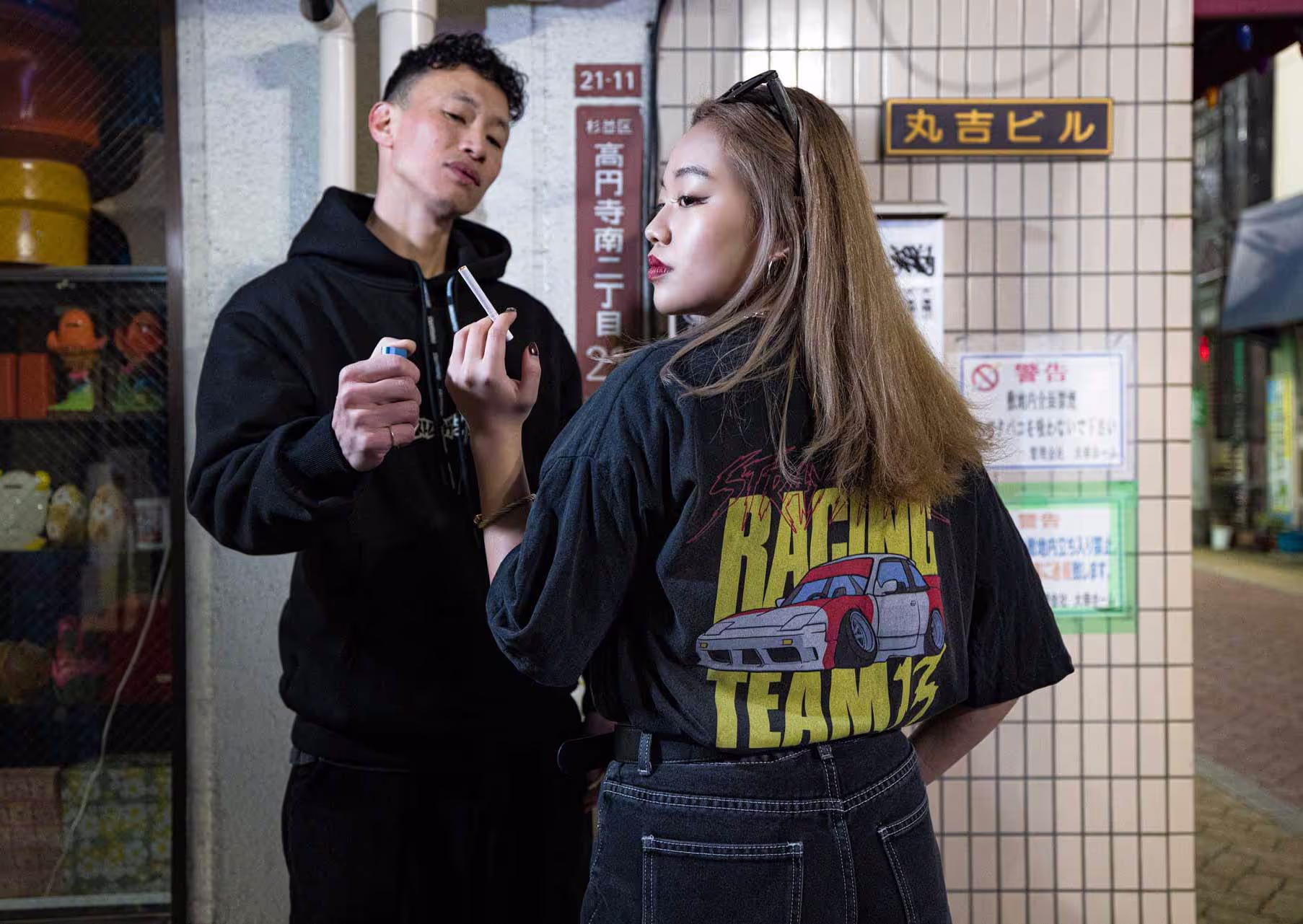 Two people in the streets of Tokyo wearing Straygang branded clothing. The most visible piece of clothing is a black wash, oversized t-shirt, showcasing the vibrant and colorful back print.