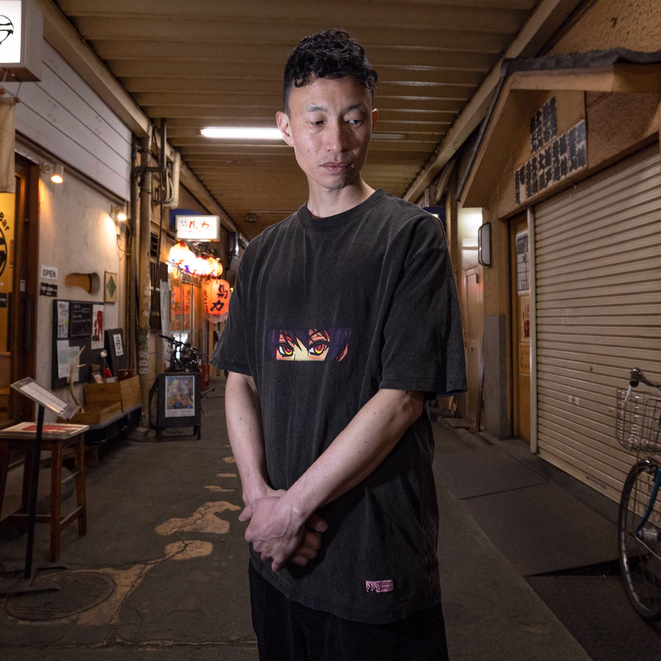 A person wearing Straygang's black wash "nostalgia" t-shirt in the dynamic streets of Tokyo. The front design features anime-style eyes in a digital print, which adds contrast to the surroundings.