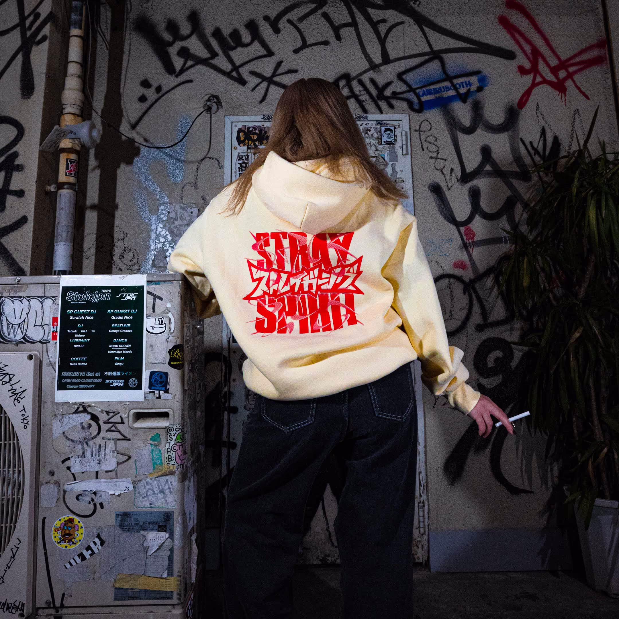 A person wearing Straygang's cream "Spirit Tag" hoodie, highlighting the back design, modeled in a Tokyo alley. The large screen-print creates a unique, visual contrast against the backdrop of the city.