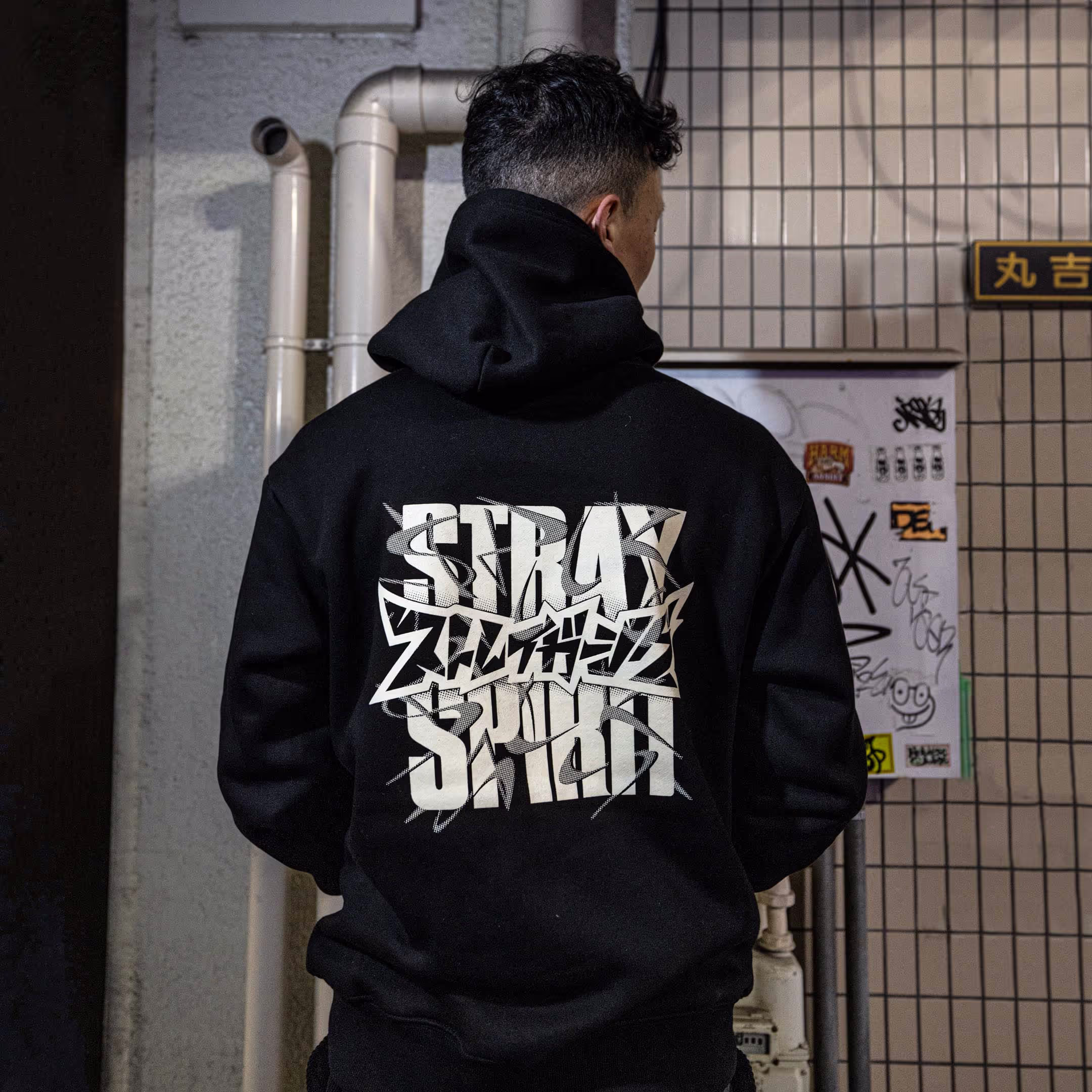 A person wearing Straygang's black "Spirit Tag" hoodie, highlighting the back design, modeled in a Tokyo alley. The large screen-print creates a unique, visual contrast against the backdrop of the city.
