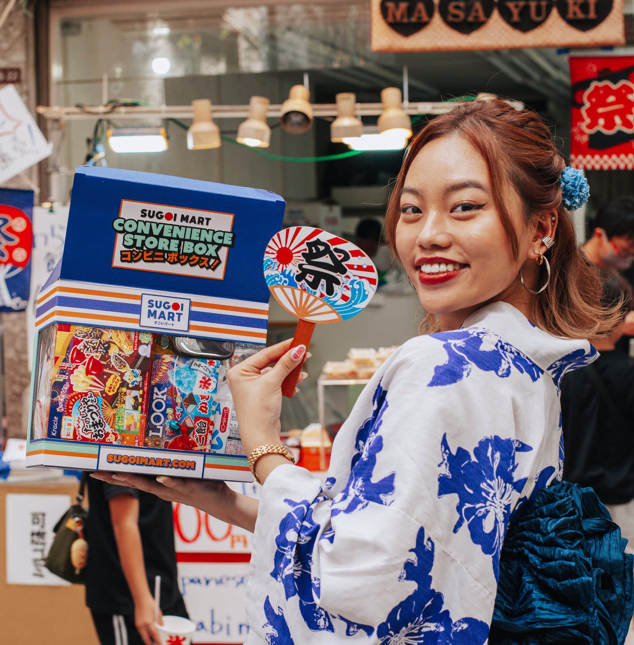 A person wearing a Japanese kimono and holding a vibrant package. The package is shaped like a mini Japanese convenience store and features artwork that further expands the "conbini" aesthetic in a colorful and cartoonish style inspired by anime and manga.