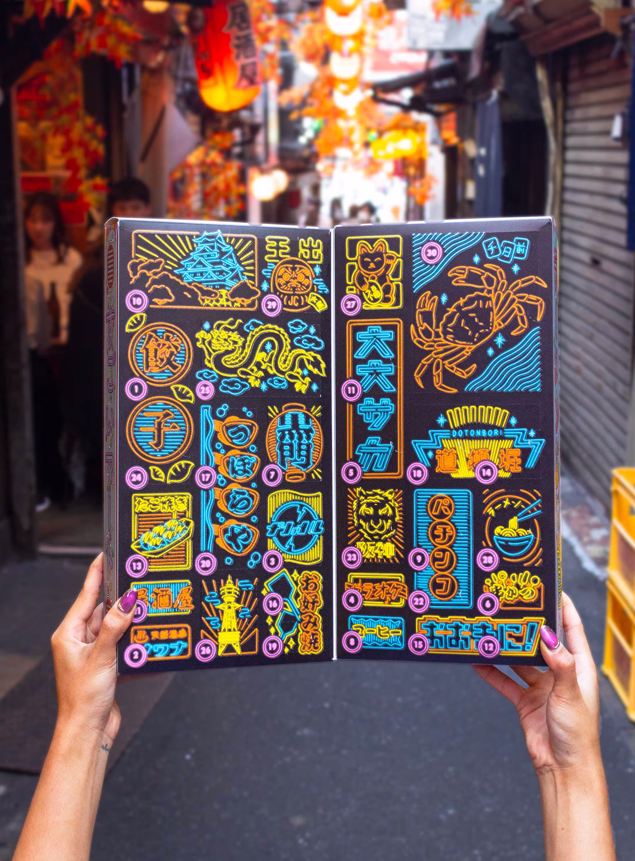 Hands holding up a vibrant and colorful package on a Japanese back alley. Showing the inside of the package. The package is an advent calendar but has a unique Japanese aesthetic to it, with all sorts of Japanese illustrations and drawings on it.